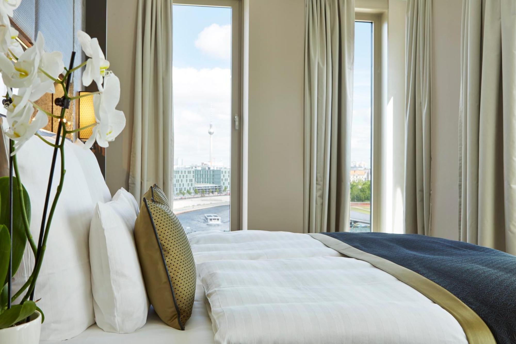 Steigenberger Hotel Am Kanzleramt Berlin Bilik gambar The photo shows a well-decorated hotel room with a large bed featuring plush white bedding and decorative pillows. There are elegant curtains framing large windows that allow natural light to enter the space. In the foreground, a vase with white orch